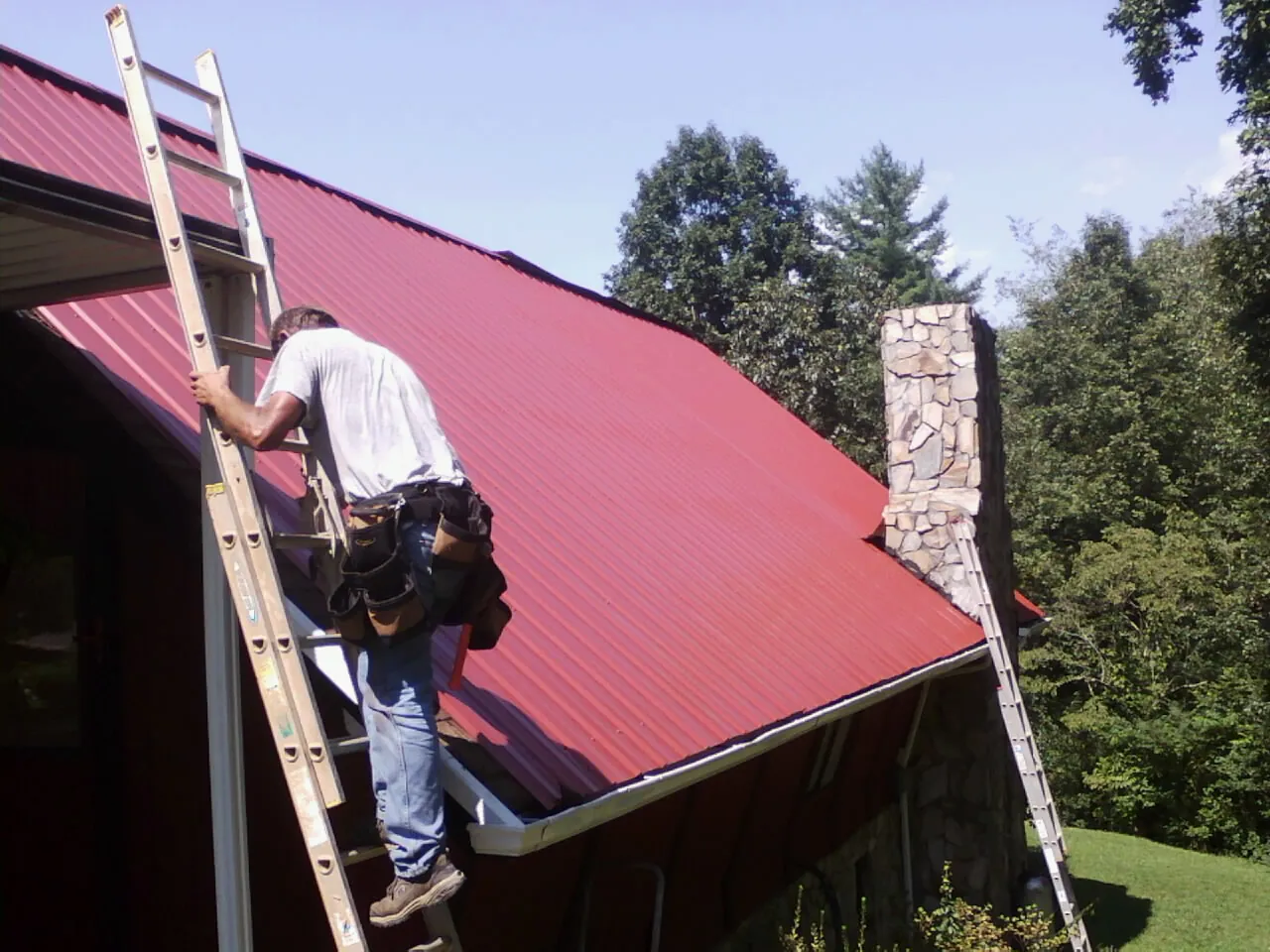 Roofer on a ladder beside a red metal roof and stone chimney — Soffit & Fascia Repair by Roofing Brothers in Gallatin